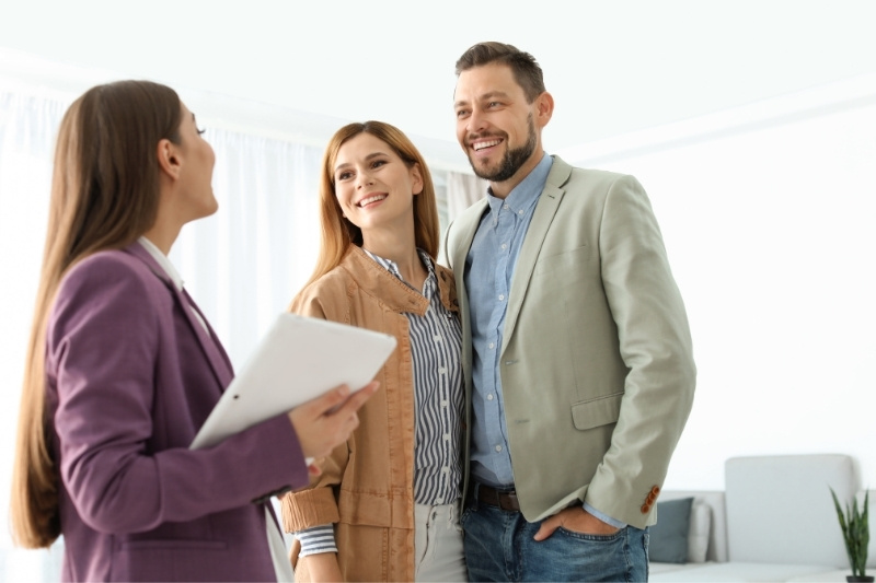 A woman holding a tablet smiles while talking to a happy couple dressed in business casual attire, standing together in a bright, modern room.