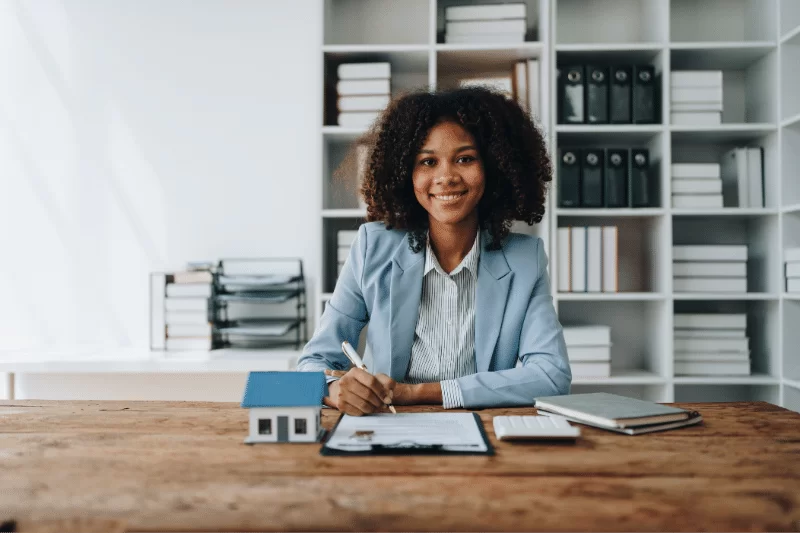 A smiling woman in a blazer sits at a desk with paperwork, a model house, a notebook, and a smartphone, in a modern office with shelves of books and binders behind her.